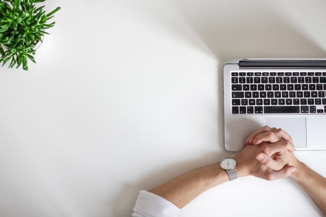 A person who is mostly out of frame is seated at a desk and clasping their hands over a laptop.