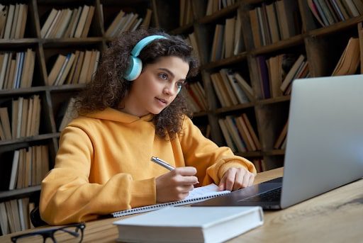 A young person in a yellow sweatshirt wears blue headphones and sits at a desk with a pen in their hand and a laptop in front of them.